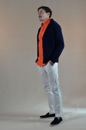 Homme de profil portant un cardigan en laine bleu marine élégant, une chemise en lin orange vif, et un chino blanc, complété par des mocassins bleu marine.